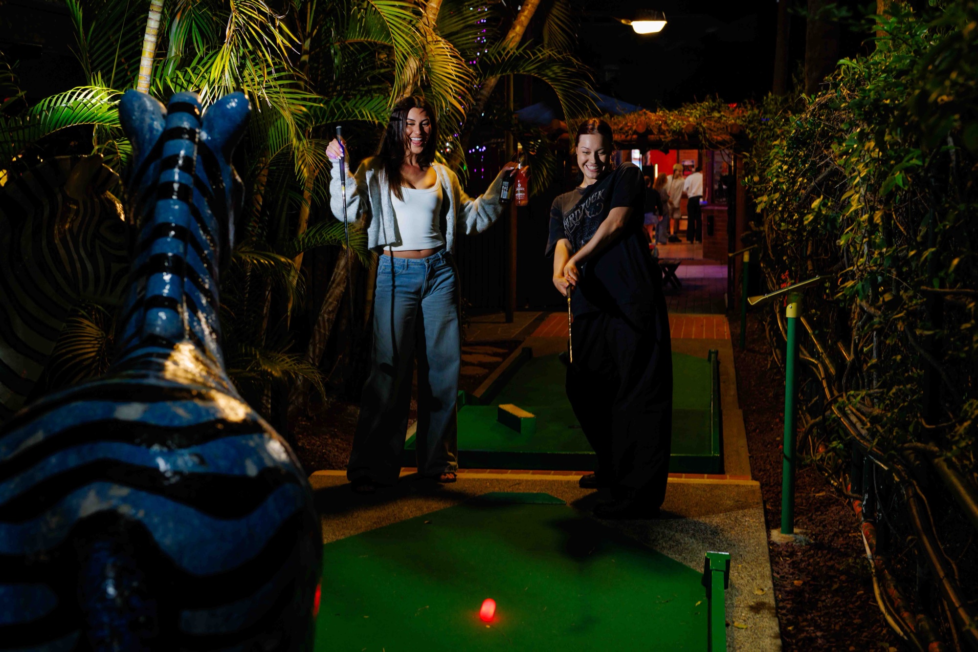 Two friends laughing over a putt on a floodlit mini golf course at night, drinks in hand